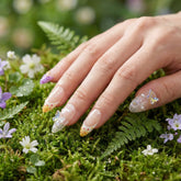 Hand with Coastal Bloom press-on nails on a background of green foliage and small flowers