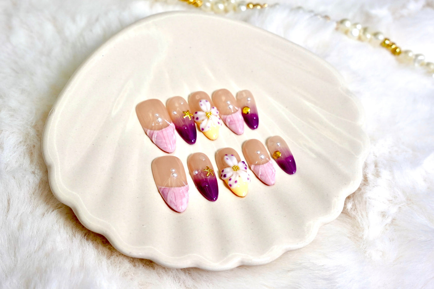 Stylet bilde av Siren's Blush press on negler for en strand-gudinne-look. De drømmende pastellneglene er presentert på et hvitt skjell-fat med en myk bakgrunn.