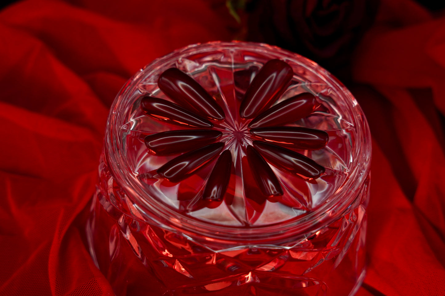 Kunstnerisk bilde av Cherry on Top-neglene arrangert i et blomstermønster på et krystallglass, med en rød satengbakgrunn.