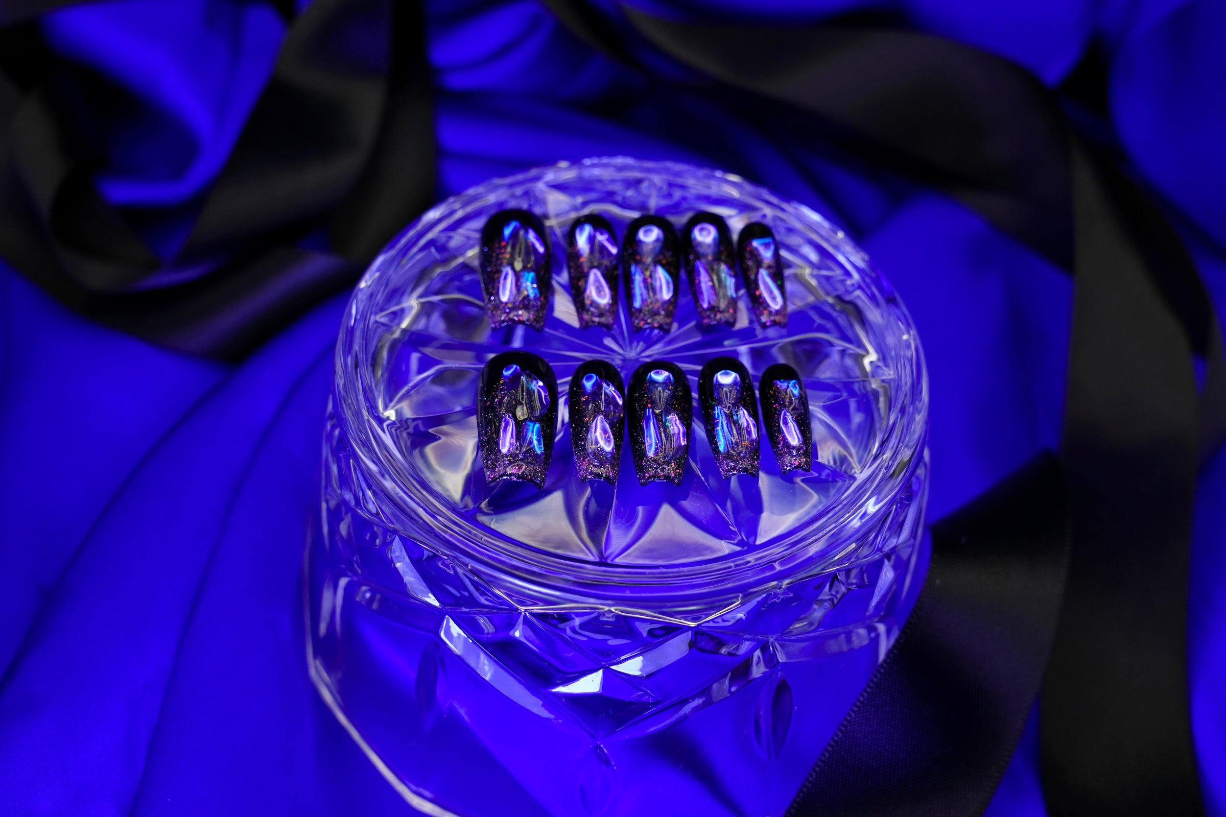 Stylet, gotisk bilde av Venom Drip press on negler. Det mørke neglesettet er dramatisk presentert på et krystallglass med en bakgrunn av blå og sort sateng.