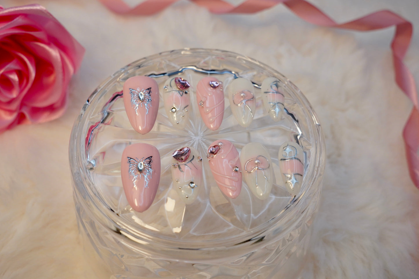 Stylet bilde av Butterfly Wish press on negler. Det delikate og drømmende neglesettet er elegant presentert på et krystallglass med en rosa rose i bakgrunnen.