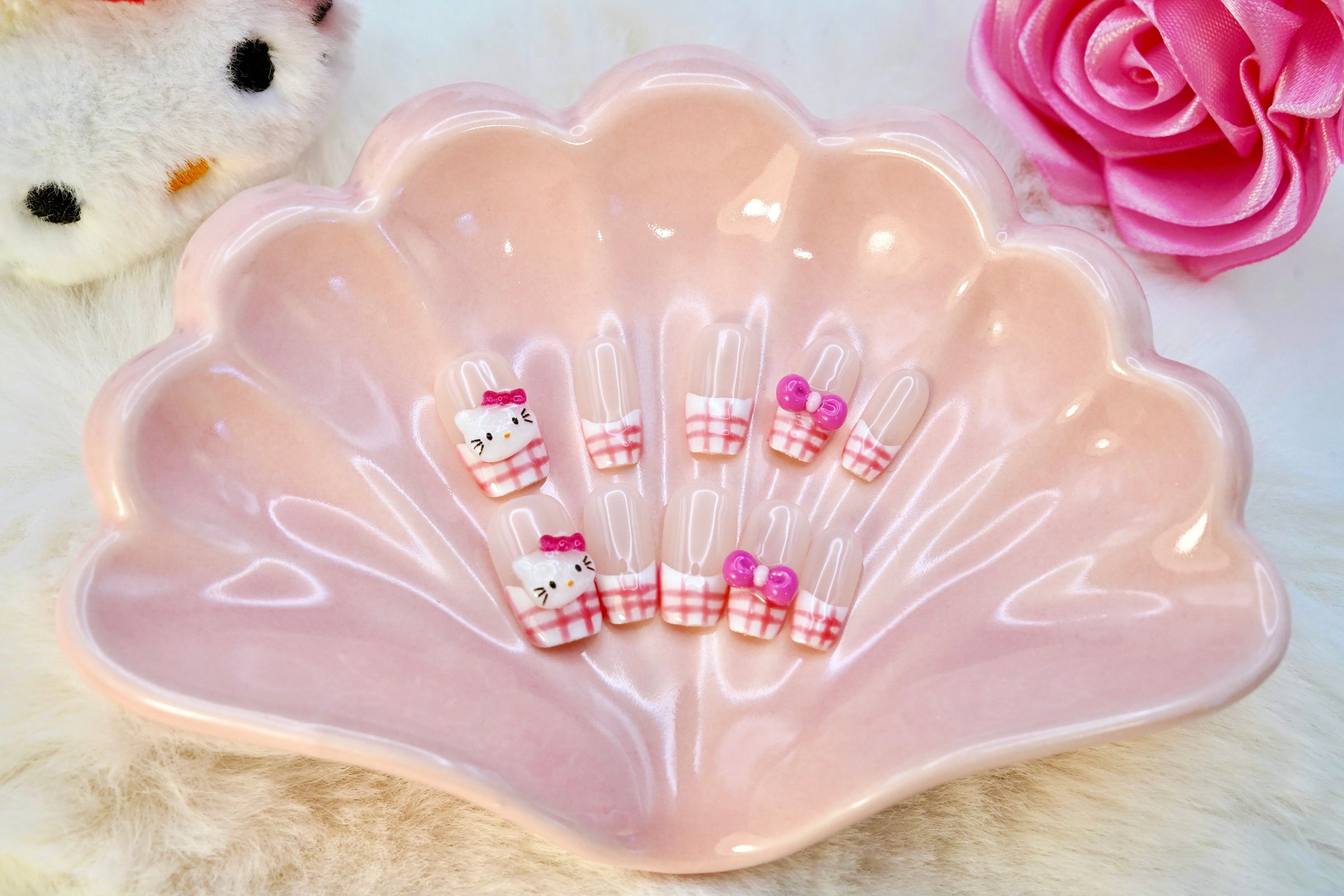 Stylet bilde av Gingham Kitty press on negler. Det søte neglesettet er presentert på et rosa skjell-fat, med en Hello Kitty-bamse i bakgrunnen.
