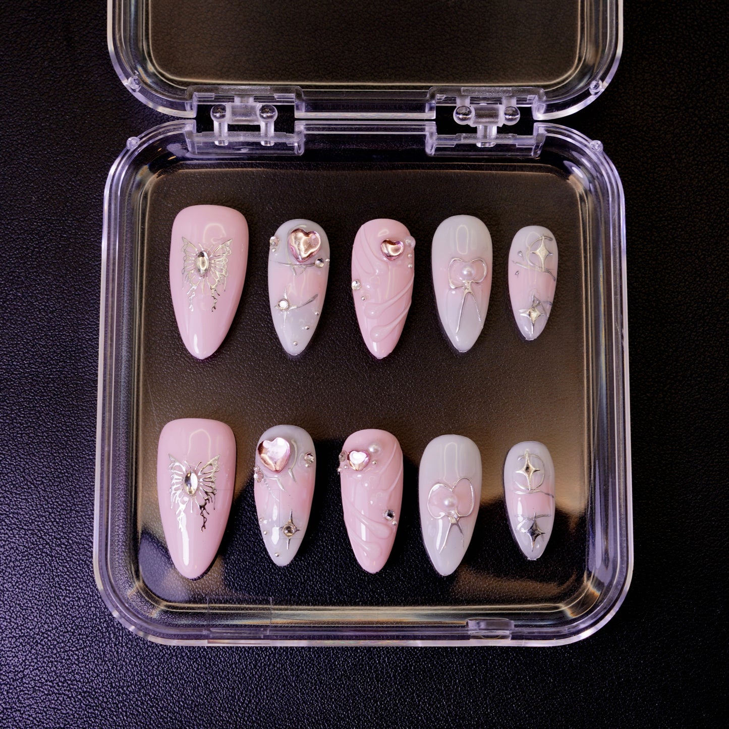 Ovenfra-bilde av Butterfly Wish press on negler i etui. Viser de mandelformede neglene med sølvfargede sommerfugler og hjerte-detaljer på en rosa og hvit base.