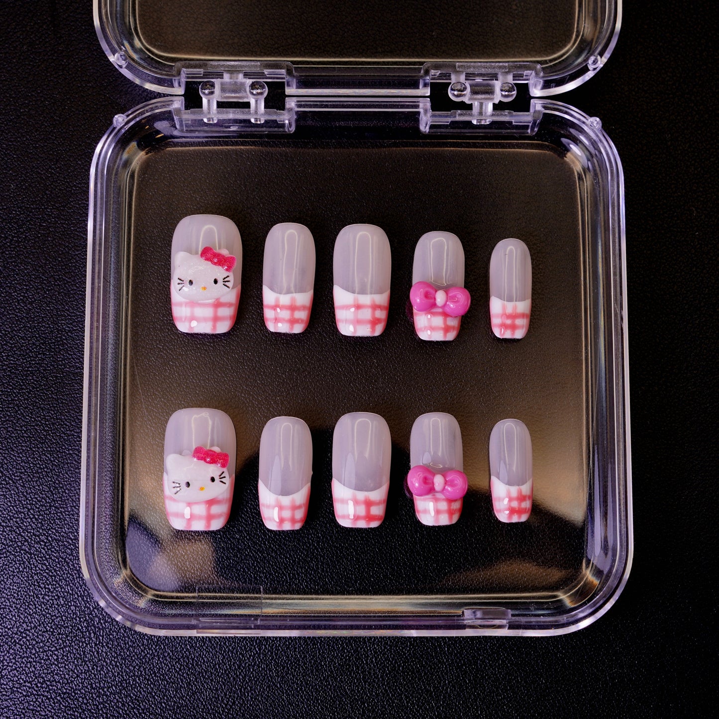 Ovenfra-bilde av Gingham Kitty press on negler i etui. Viser de firkantede neglene med rosa gingham-mønster, Hello Kitty-ansikter og 3D-sløyfer.