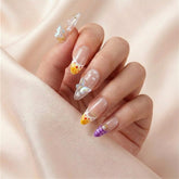 Hånd på lyserosa silkestoff med Coastal Bloom press on negler — håndlaget, gjenbrukbare almond-negler med 3D iridescent sommerfugler, perler og oransje-gul French tip.