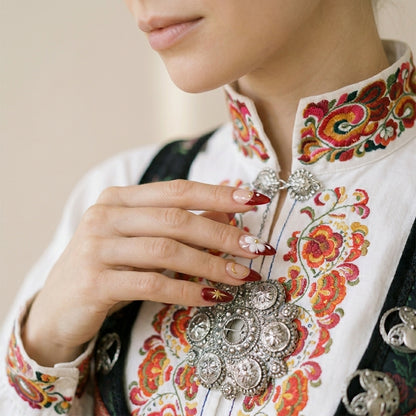 Hånd hviler på bunad med Telemark-broderier og sølv-sølje — Rosemaling Royalty press on negler med vinrød ombré, hvite 3D blomster og sølje-inspirert gullstjerne.