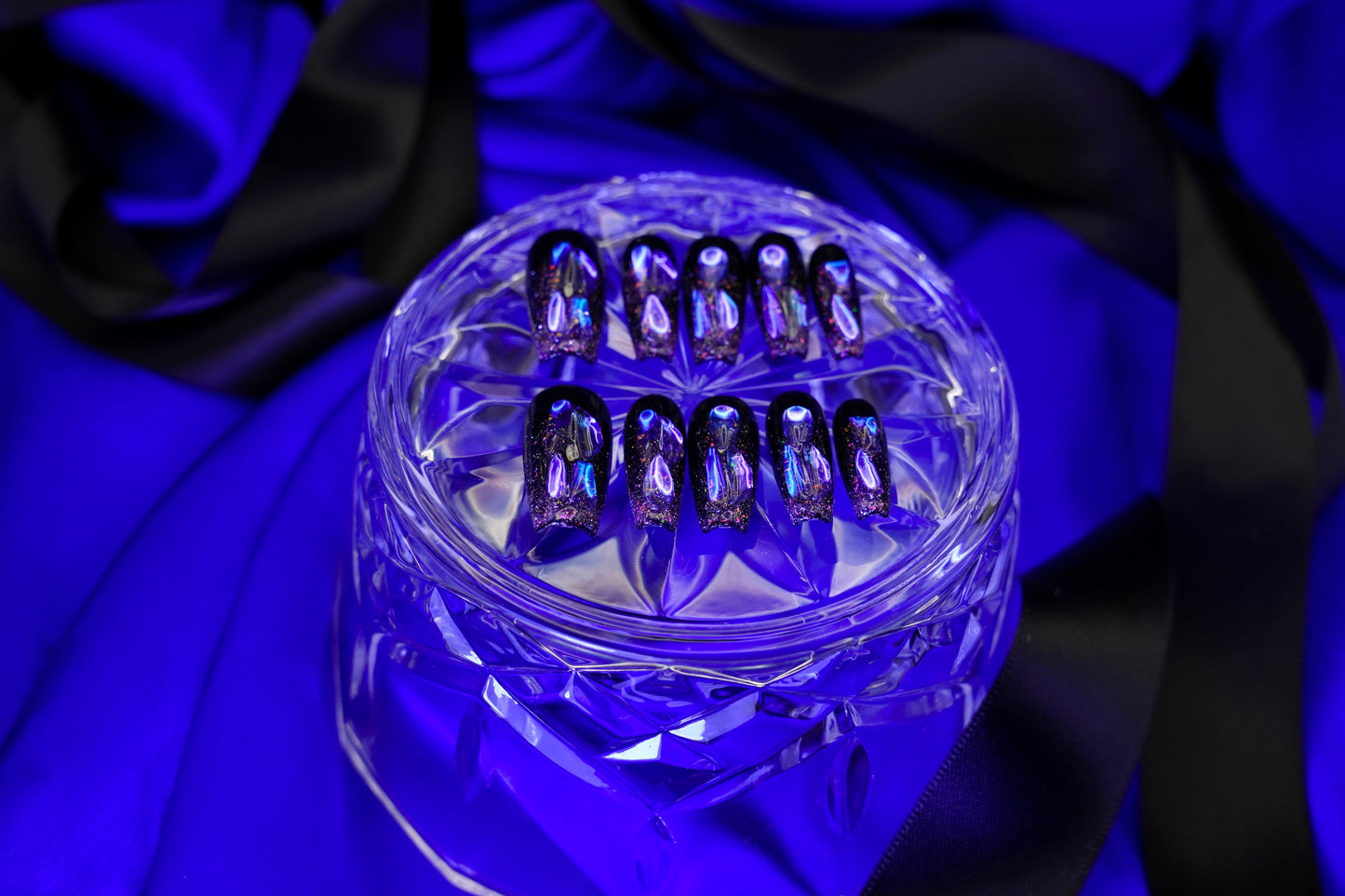 Stylet, gotisk bilde av Venom Drip press on negler. Det mørke neglesettet er dramatisk presentert på et krystallglass med en bakgrunn av blå og sort sateng.