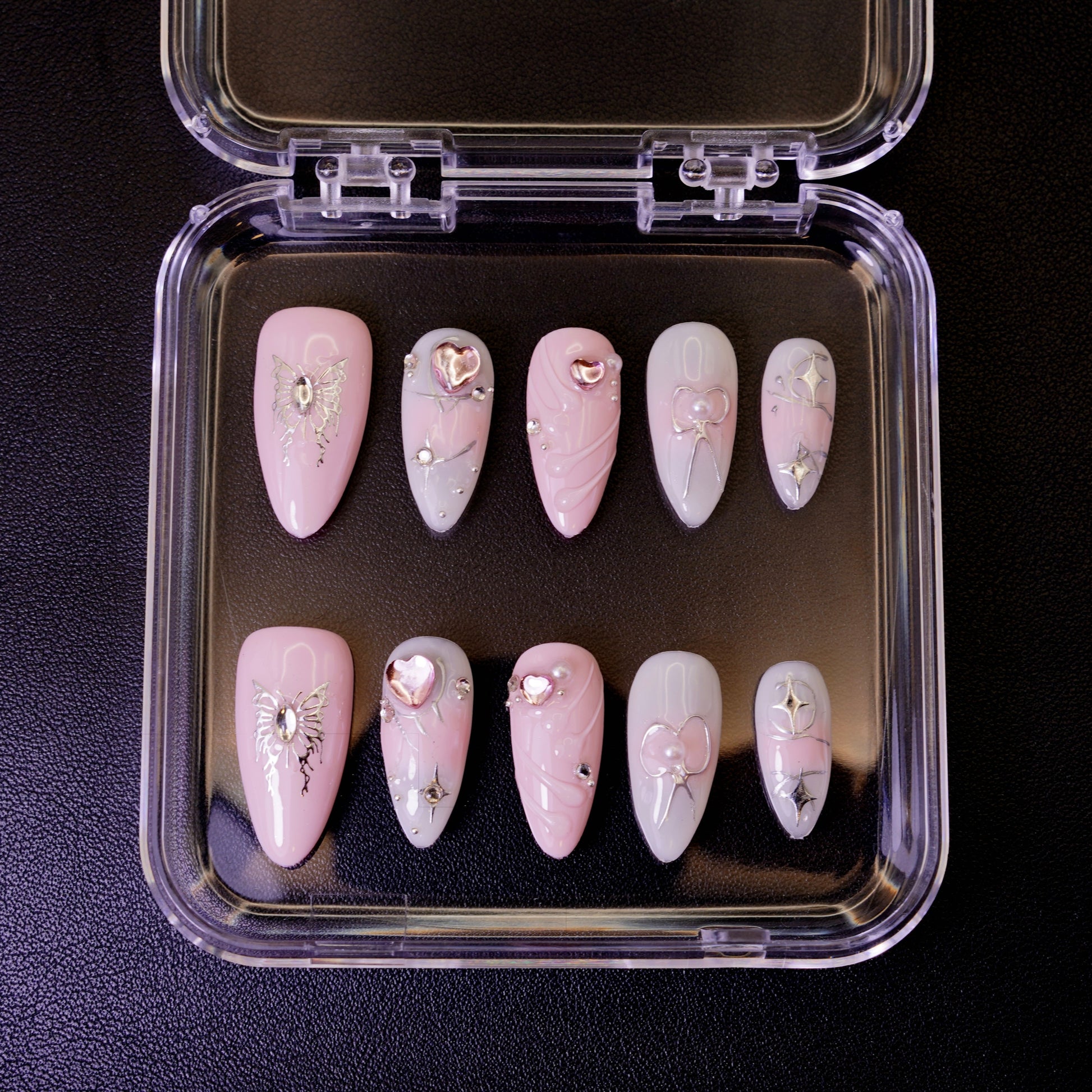 Ovenfra-bilde av Butterfly Wish press on negler i etui. Viser de mandelformede neglene med sølvfargede sommerfugler og hjerte-detaljer på en rosa og hvit base.