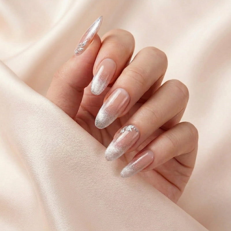 Hånd på lyserosa silkestoff med Angel Glaze press on negler — håndlaget, gjenbrukbare almond-negler med nude-rosa til sølv-glitter ombré og 3D sølv-blad accent.