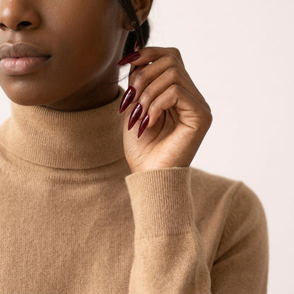 Kvinne i kamelfarget turtleneck hever hånden med Cherry on Top press on negler — lange stiletto-negler i glossy kirsebærrød chrome-finish.