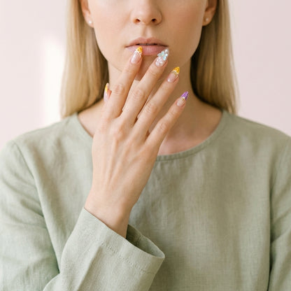 Hånd løftet mot munnen i lin-bluse i salviegrønt — Coastal Bloom press on negler i almond-form med gul French tip, perler, sommerfugl- og sjøstjerne-charms.