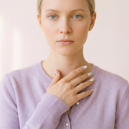 Nærbilde av kvinne i lys lilla kardigan som hviler hånden på brystet — Cotton Candy press on negler i pastell chrome almond-form med rosa, grønn og gul shimmer.