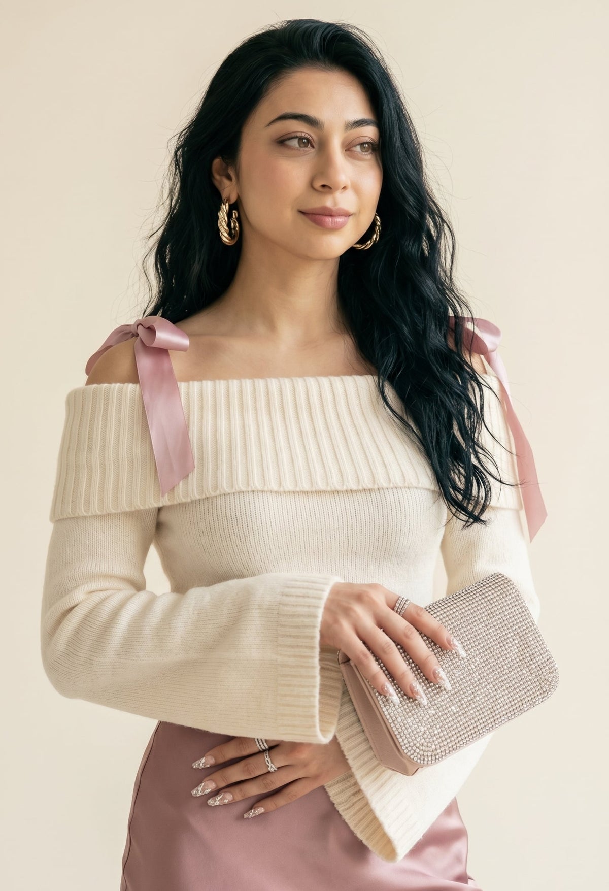 Editorial portrait — mørkhåret kvinne med store gullringer, off-shoulder strikkegenser med rosa sløyfer og sateng nederdel, holder krystall-clutch med nude French-inspired negler.