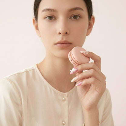 Kvinne holder rosa macaron opp mot ansiktet, viser Kawaii Crisis press on negler — almond-form med 3D Hello Kitty charms, rosa hjerter og myke pastellfarger.