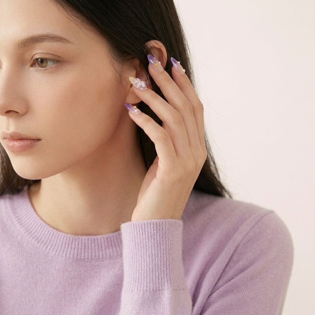 Detalj av hånd i lyselilla genser som rører håret — Siren's Blush press on negler i almond-form med lilla ombré, 3D hvit blomst- og perle-detaljer.