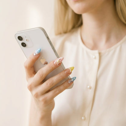 Hånd holder hvit iPhone i cremefarget bluse — Starstruck in Waikiki løsnegler i pastell almond-form med sommerfugl-charm, stjerner og gule-blå French tips.