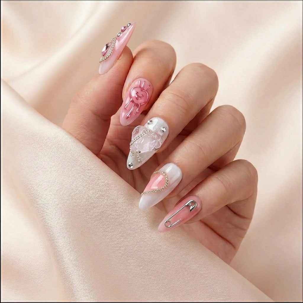 Styled hånd på rosa silkestoff med Pretty in Pain press on negler fra The Y2K Edit — almond-form med 3D rosa bamse, sølv hjerte, sikkerhetsnål og krystall-detaljer.